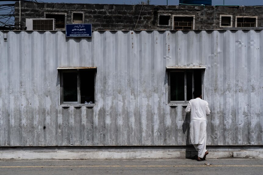 A man stands at a window of a corrugated iron building with a sign reading 'immigration' 