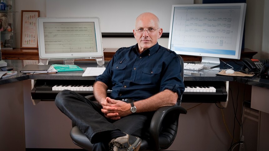Nigel Westlake sits facing the camera in front of two computer monitors with sheet music and a keyboard.