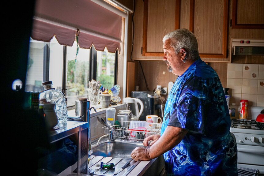 An older man stands in his kitchen and looks out the window.