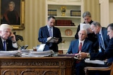 Donald Trump sits at a desk on a phone while five men stand and sit on the other side of the desk
