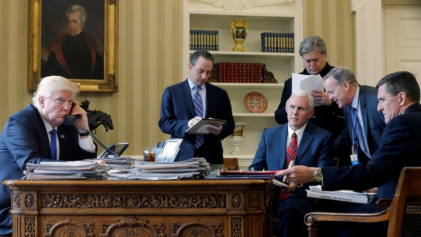 Donald Trump sits at a desk on a phone while five men stand and sit on the other side of the desk