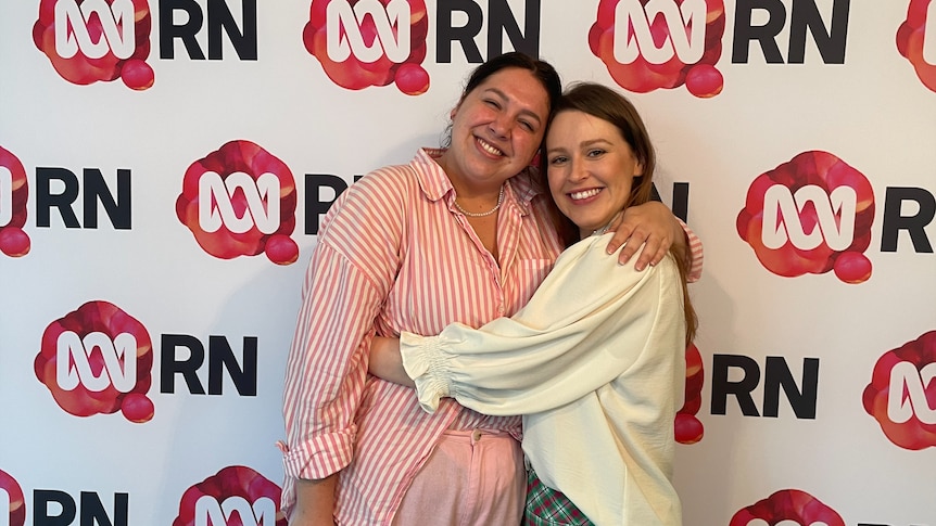 two women hug and smile into the camera against a photo wall with RN Branding