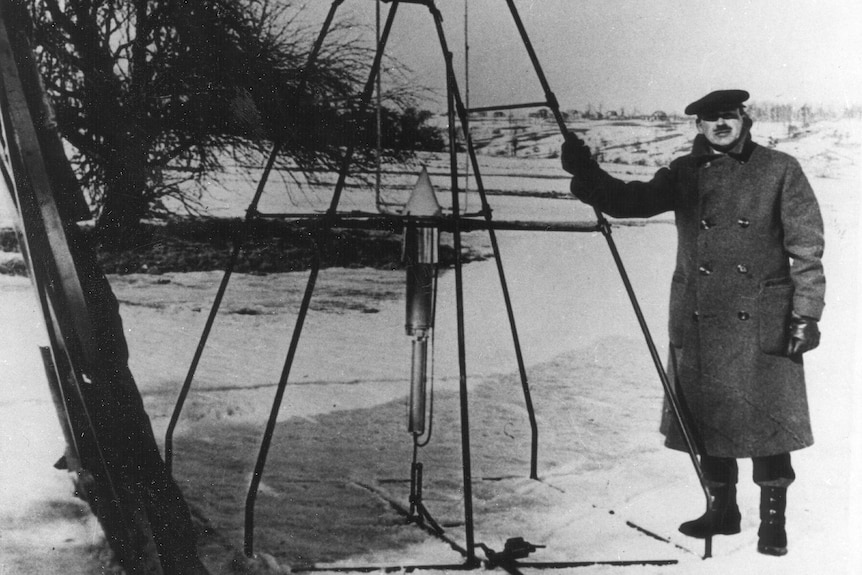 Man stands next to tall metal structure with small rocket at the top.