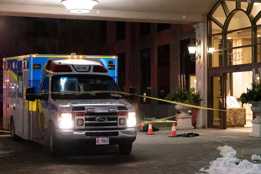 An ambulance is parked behind police tape next to the open door of an apartment building at night. 