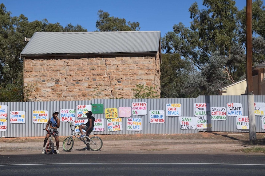 White sheets of paper with phrases demanding water painted in colours scattered on a fence. two boys on bikes sit in front