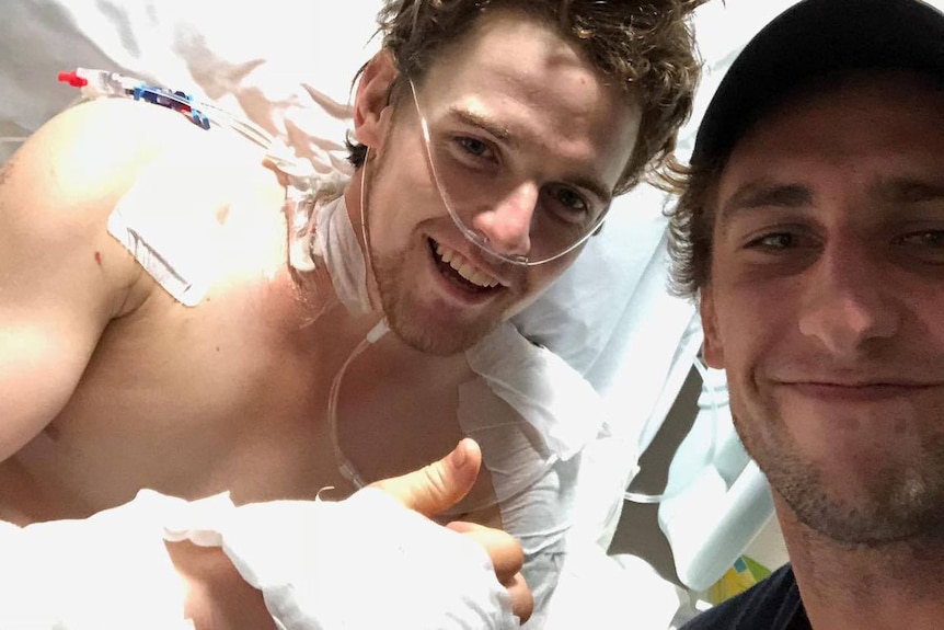 Smiling man in bandages lying in hospital bed with a thumb up with man on right taking the selfies with smiling face