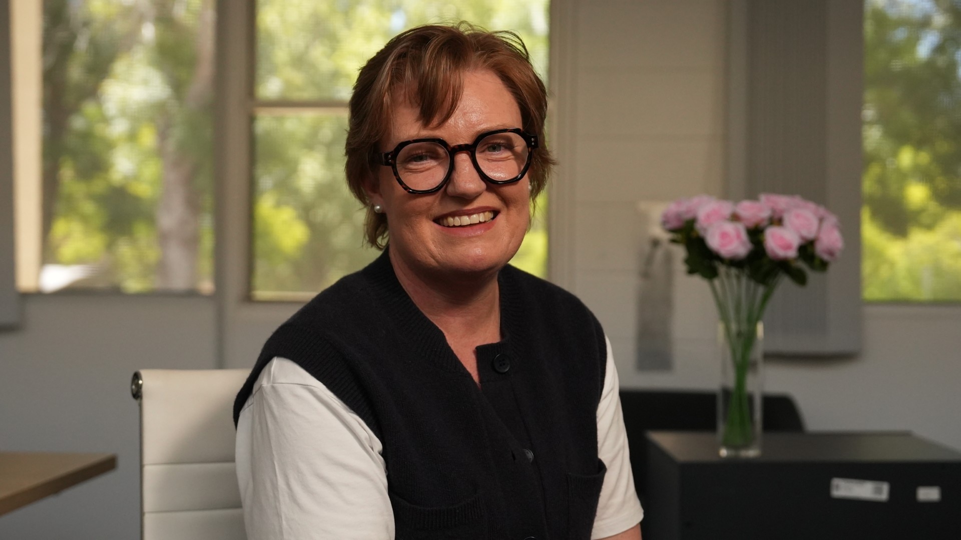 Woman wearing glasses smiling at the camera with flowers in the background