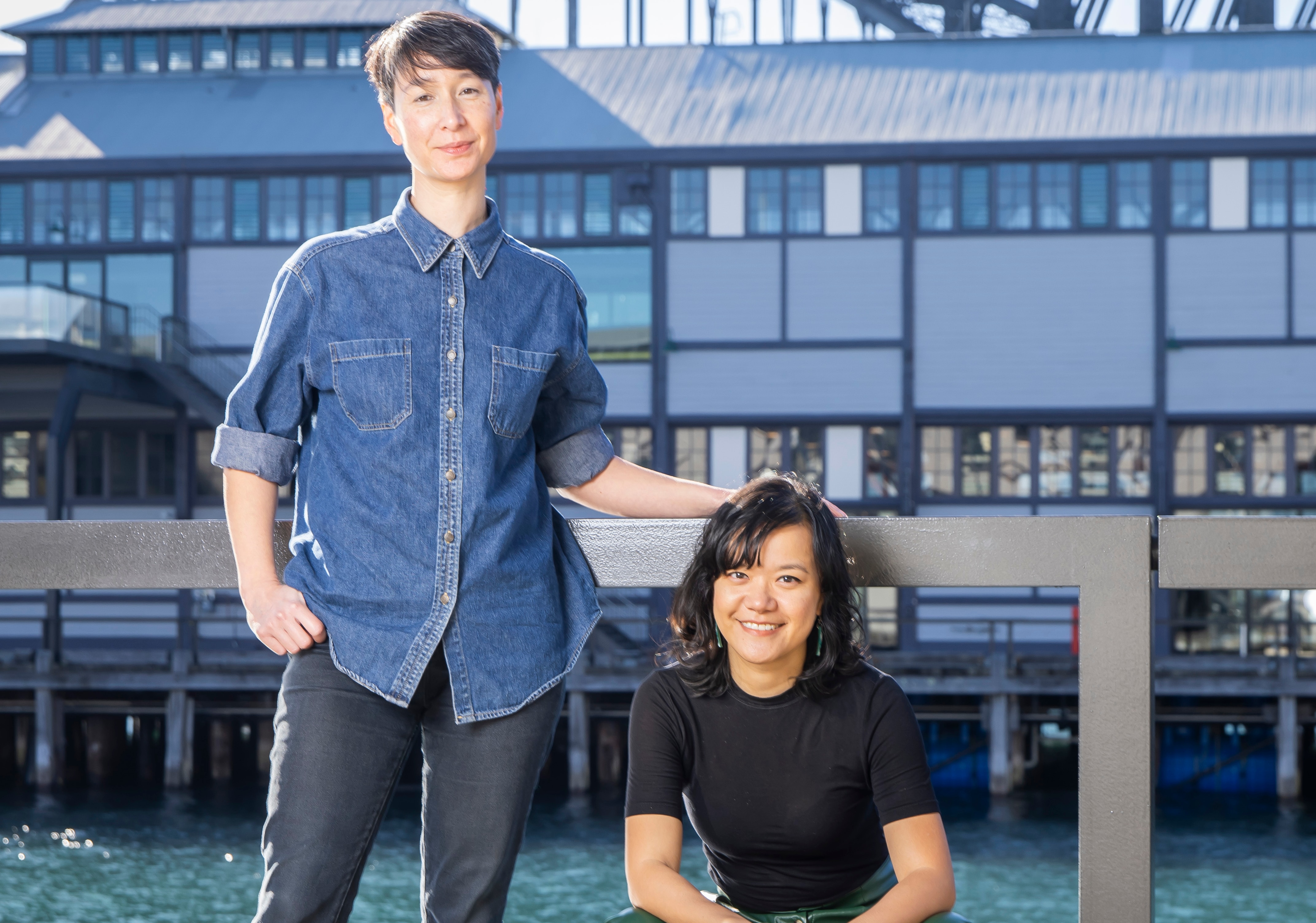 Two 30-something Asian Australian women, one standing, the other sitting, are on a pier by the harbour on a bright day, smiling