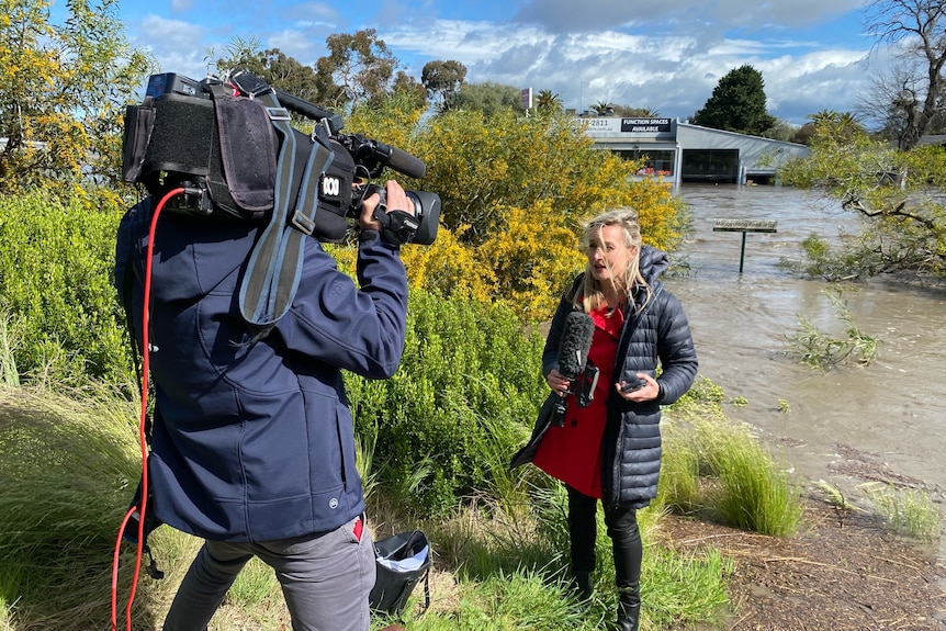 Cameraman filming woman holding microphone standing on bank of flooded river.