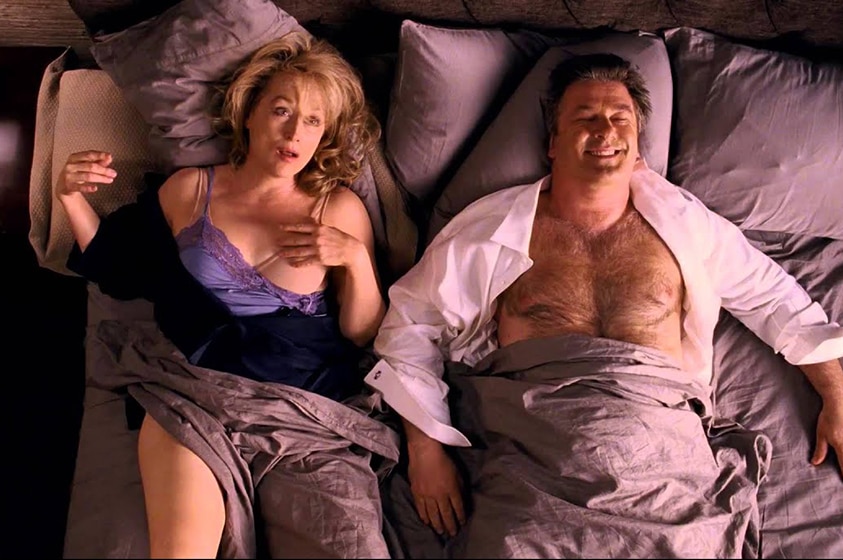 A middle aged man and woman with shock but pleased expressions lay semi-undressed together in messy bed.