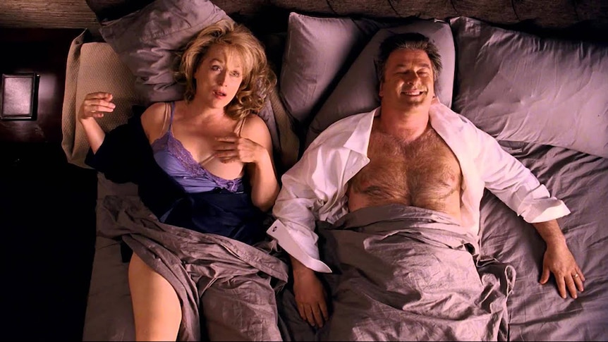 A middle aged man and woman with shock but pleased expressions lay semi-undressed together in messy bed.