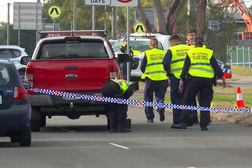 Four police officers in high vis vests stand near a red ute parked in the middle of a suburban street.