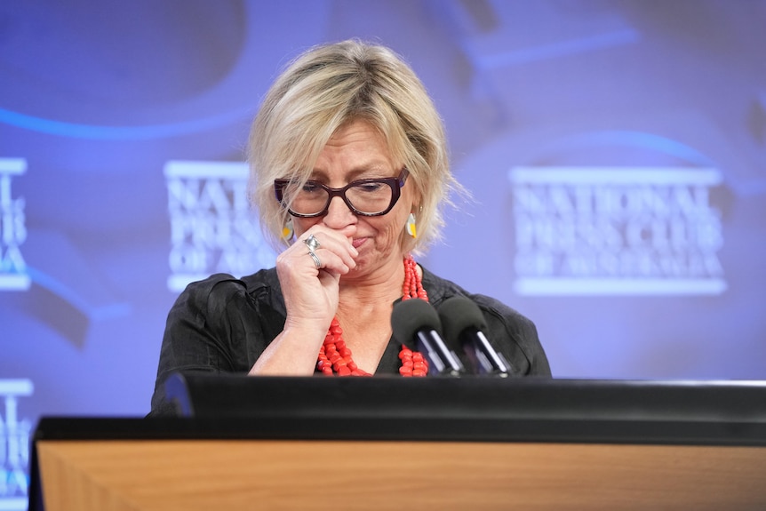 A white woman, trying not to cry. She's standing at a lectern in front of a purple background.