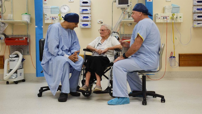 Two doctors wear blue uniforms and caps and sit next to an old woman. They are surrounded by equipment in a clinical space