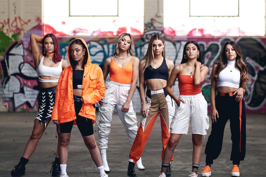 Six women standing in a row wearing orange, black and white urban street dancing costumes 