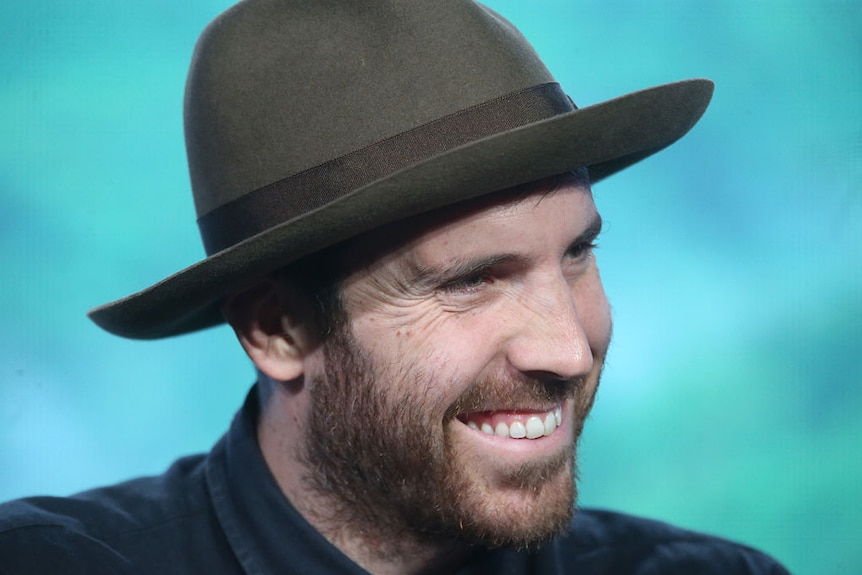 Close-up of director Thomas M Wright wearing a brownish felt hat and smiling, at a panel at a conference indoors.
