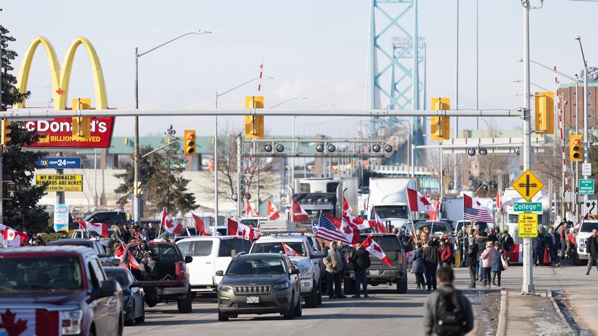 Cars and trucks with Canadian and American flags are parked on a four lane highway with a bridge in the background.