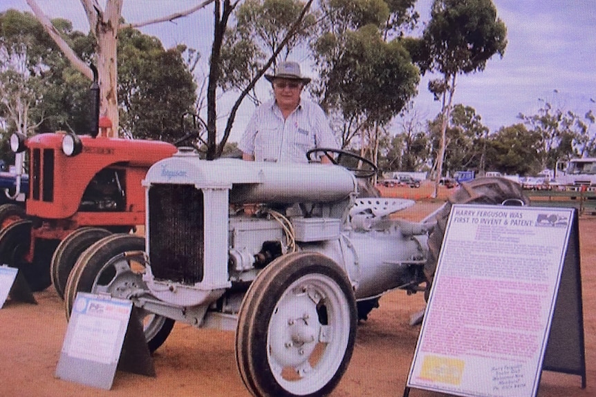 A man in a hat standing behind a classic car in a rural setting