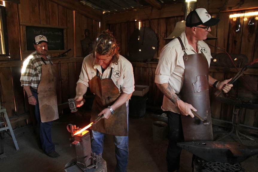 Three men in a wooden shack forging knives, all wear leaterh aprons, two wear black and cream hats.