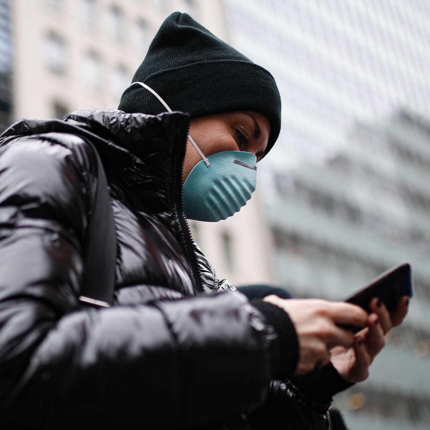 A woman in a black jacket wearing a dark beanie and a green face mask looks at her mobile phone.
