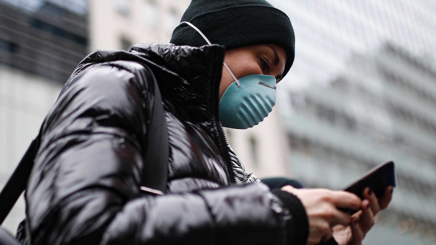 A woman in a black jacket wearing a dark beanie and a green face mask looks at her mobile phone.