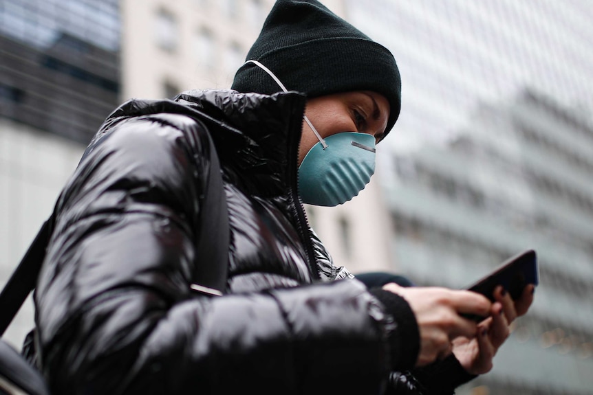 A woman in a black jacket wearing a dark beanie and a green face mask looks at her mobile phone.