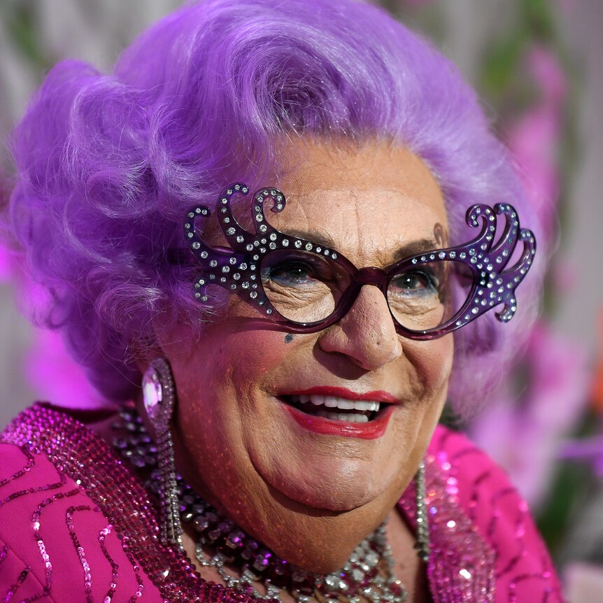Barry Humphries in drag as Dame Edna Everage, who has large glasses, bright purple hair and loud makeup.