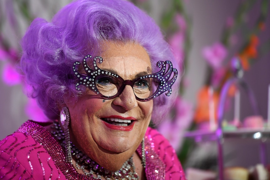 Barry Humphries in drag as Dame Edna Everage, who has large glasses, bright purple hair and loud makeup.