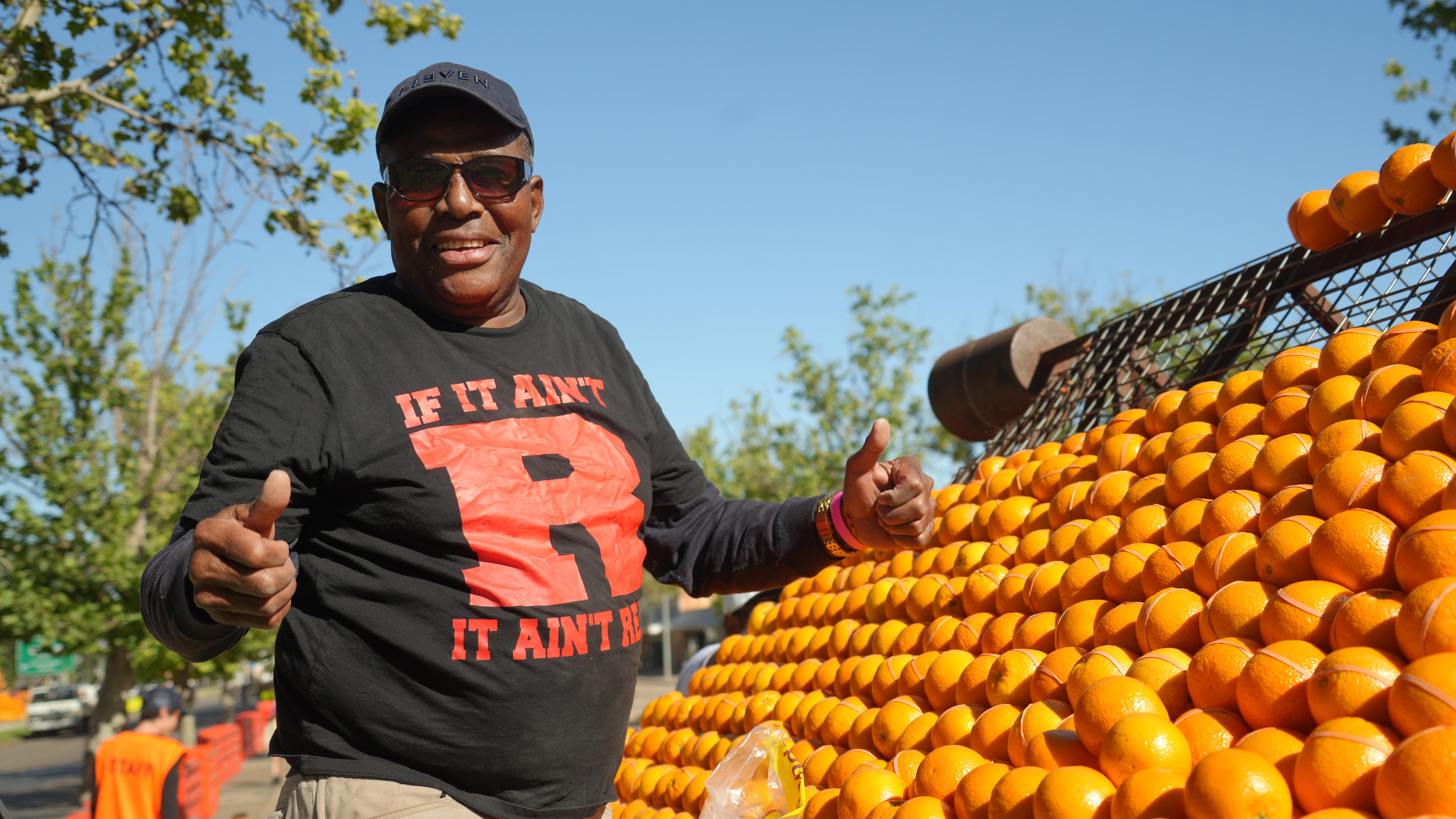 A smiling man in a cap and sunglasses stands next to a frame bearing many oranges.