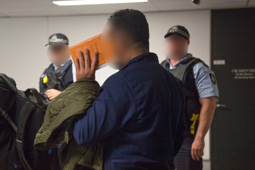 Two AFP officers with their faces blurred stand next to a man covering his face with a book