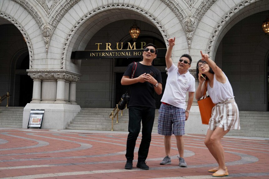 A group of people point to a person in a window of a building across the street while standing in front of the Trump Hotel