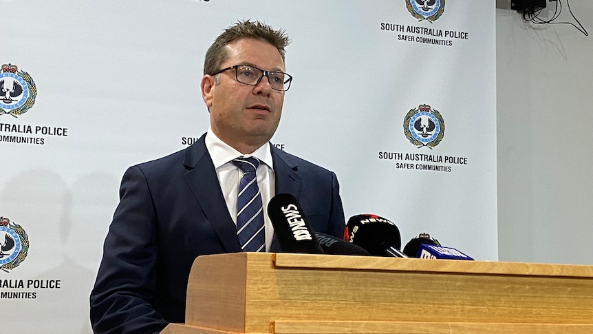 A man wearing a suit and tie stands at a lectern with an SA Police banner behind him