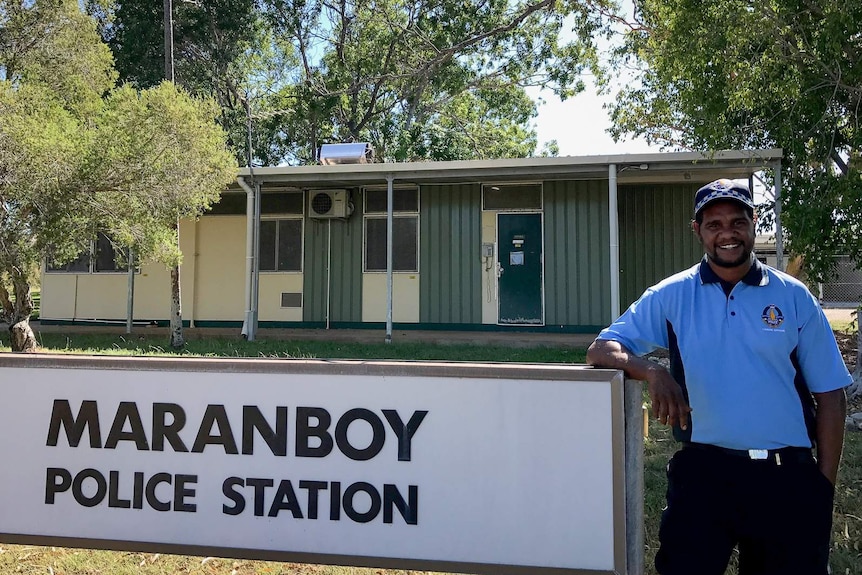 An Indigenous man wearing a blue police liaison officer's uniform stands in front of a small, single-level building.