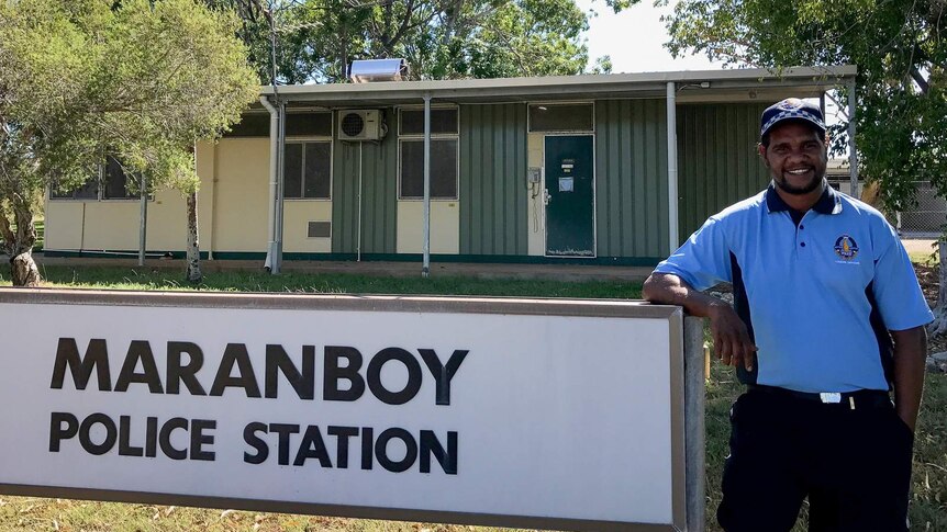 An Indigenous man wearing a blue police liaison officer's uniform stands in front of a small, single-level building.