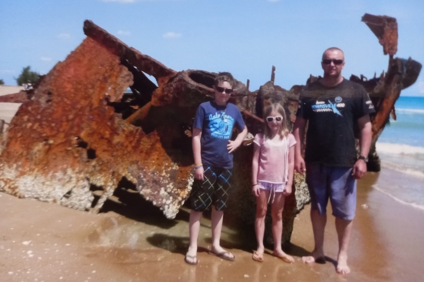A man, a girl and boy stand in front of a ship wreck on a beach.