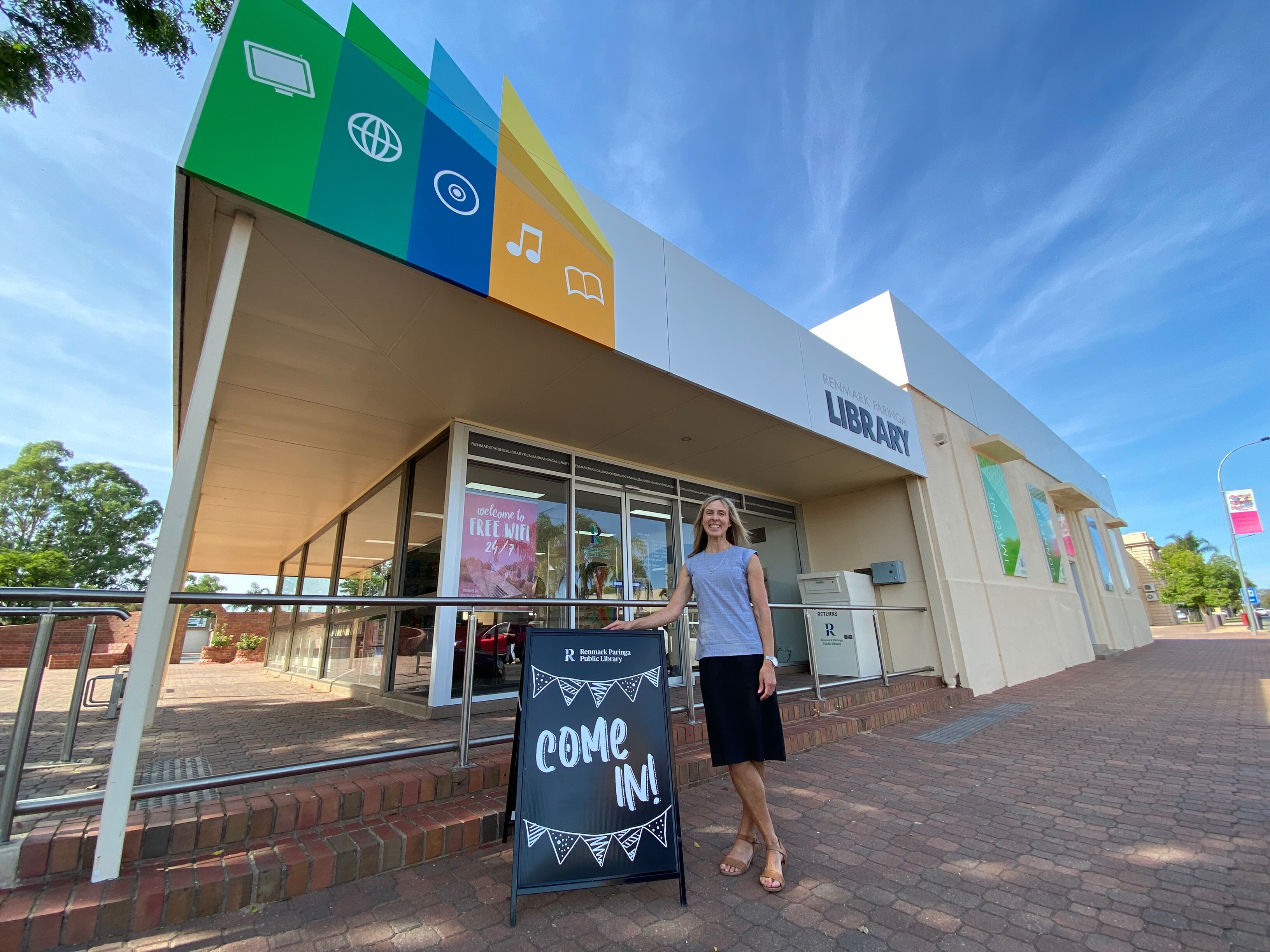 A tall, blonde white woman standing outside a building that says library and a sign that says come in