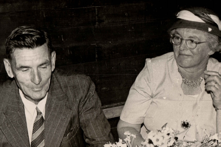 A black and white photo of a man in a suit next to a woman with a hat on. 