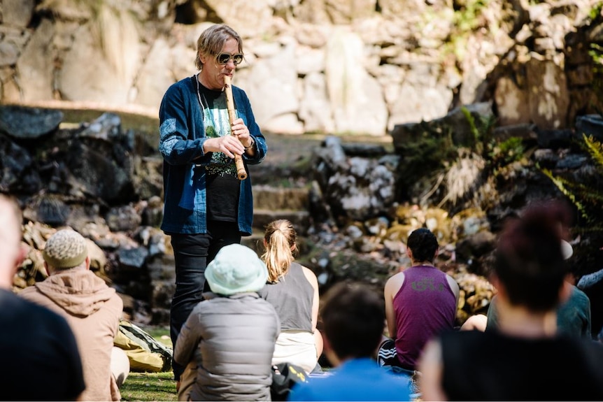 An older man wearing sunglasses and playing a bamboo flute stands in front of a group sitting on the ground in a rocky knoll