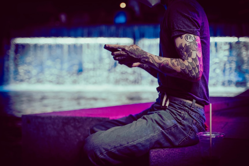 A man with heavily tattooed arms texts on his phone while sitting on a bench overlooking a river