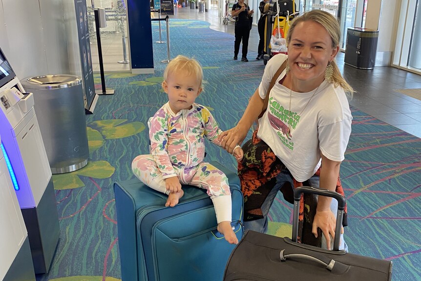 A blonde woman smiles at the camera, her small child sitting on top of suitcases at the airport
