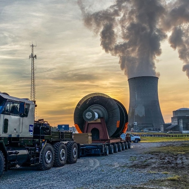 A large truck with a yellow oversized vehicle sticker has machinery on the back with two large steam stacks in the background