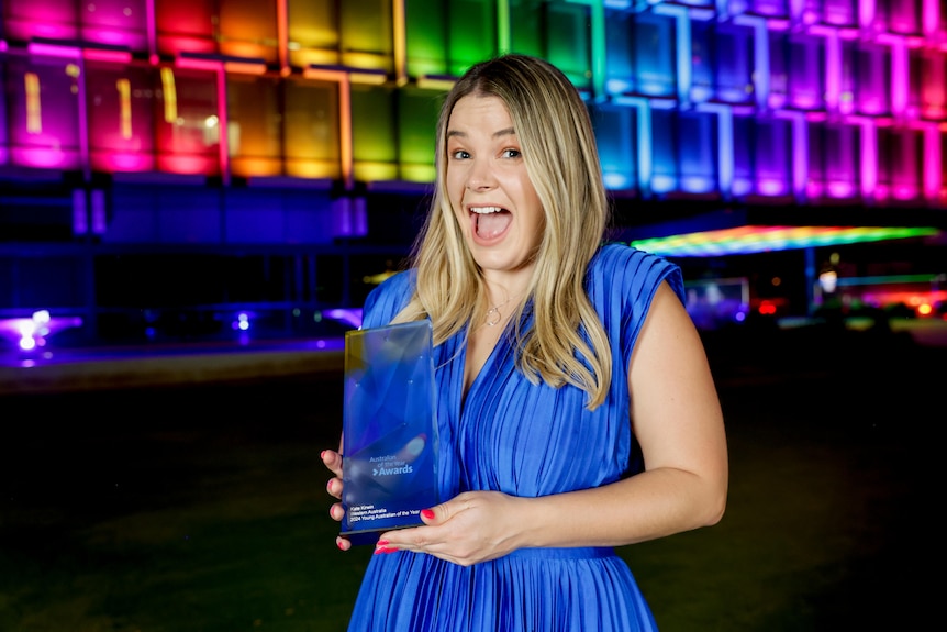 A woman in a blue dress holds a glass award in front of a building lit up rainbow.