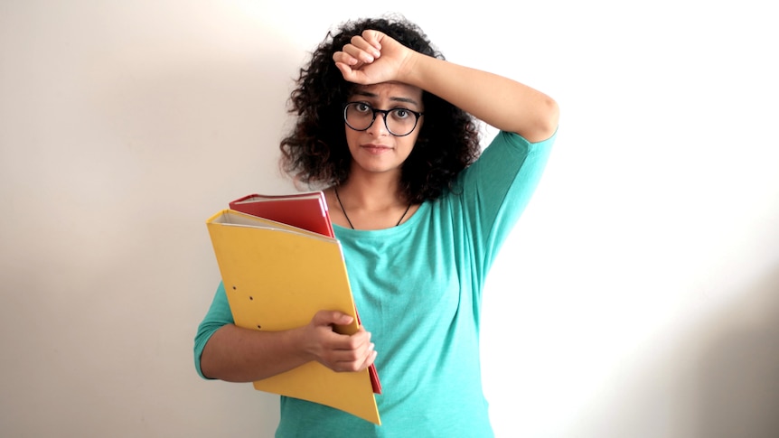 A woman holds her hand to her head while holding ring binders, for story about working multiple jobs.