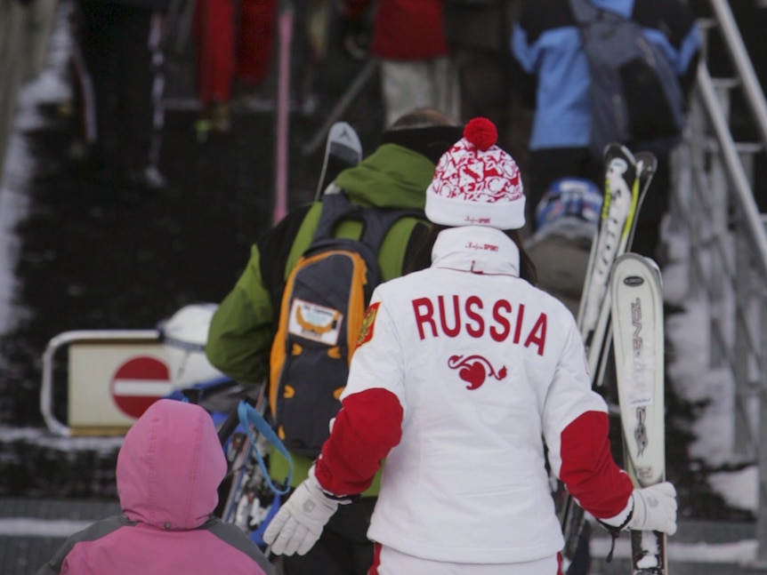 Les gens en tenue de ski sont vus de derrière, montent un escalier.  L'un porte un costume blanc avec une bordure rouge et la RUSSIE en lettres rouges