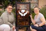 Two women kneel either side of a pantry box filled with food that says 'Neighbours helping neighbours'.