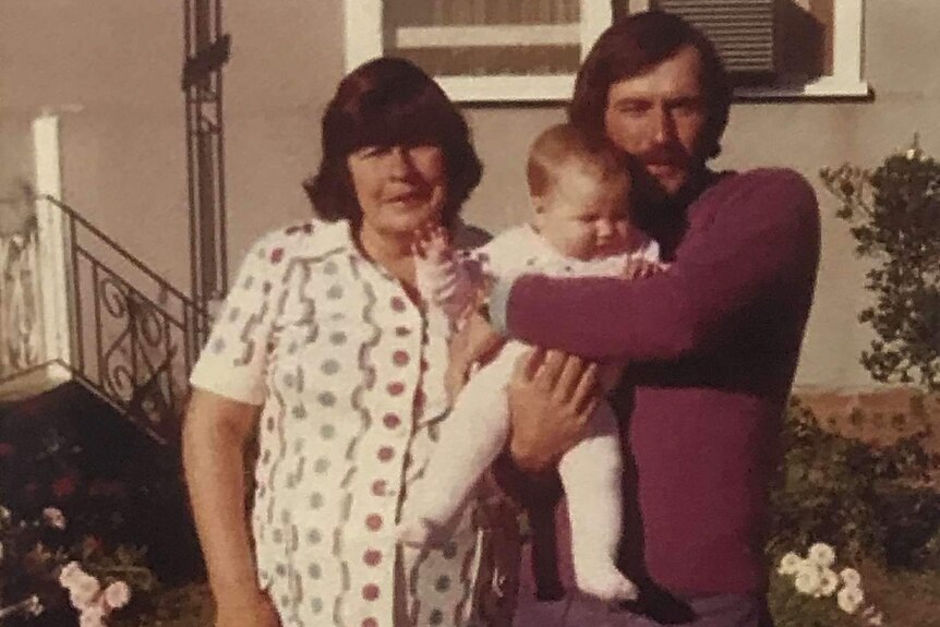 An older woman standing with a younger man who is holding a small baby on the front lawn of a house.