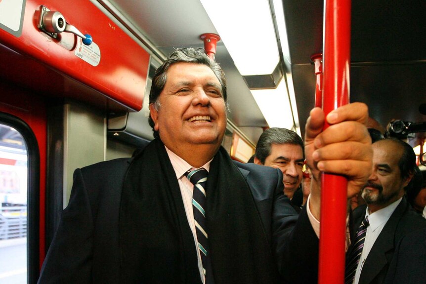 Alan Garcia smiles wearing a tie and holding onto a red pole as he rides the train.