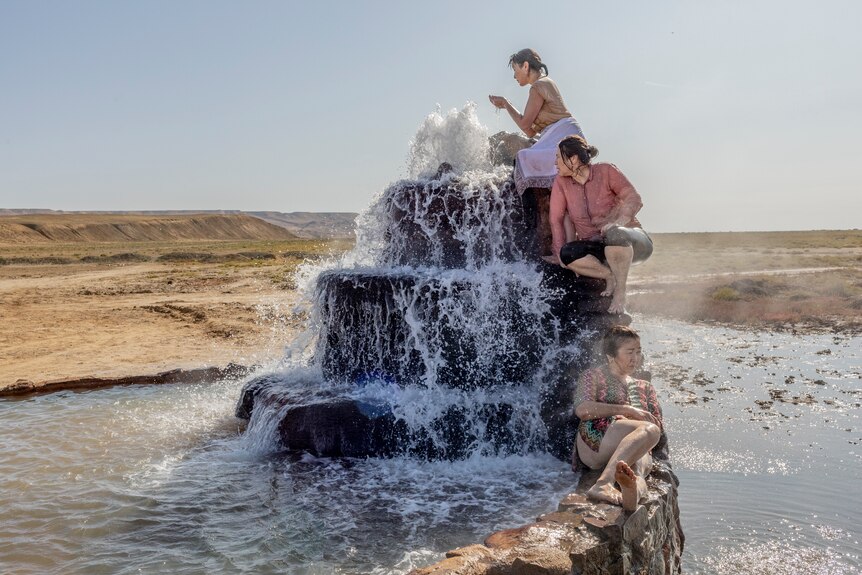 A group of women sit and lie on a rock wall fountain in a grass plains environment.