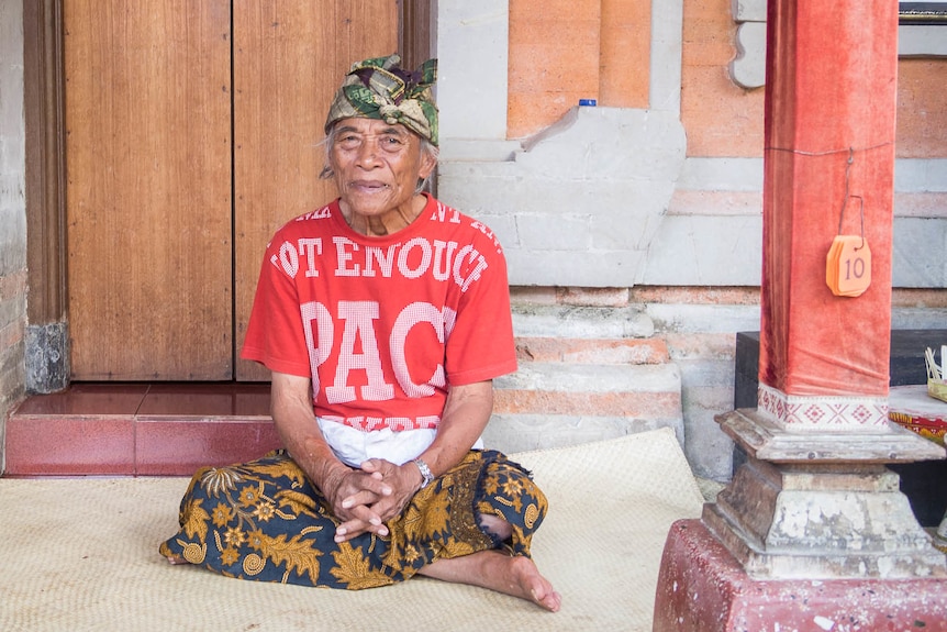 An elderly man in traditional Balinese dress sits cross legged on the floor smiling.