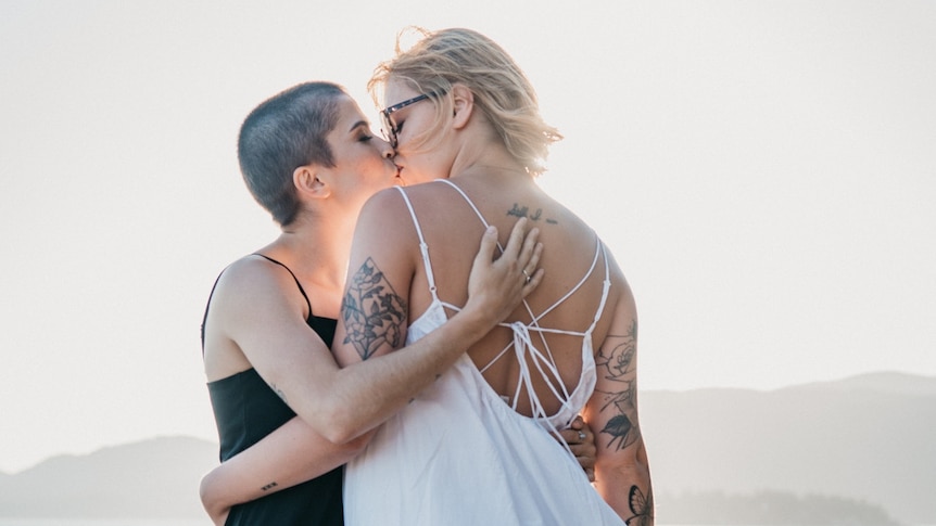 Two young women kiss on a beach, wearing dresses, wedding rings and carrying flowers.